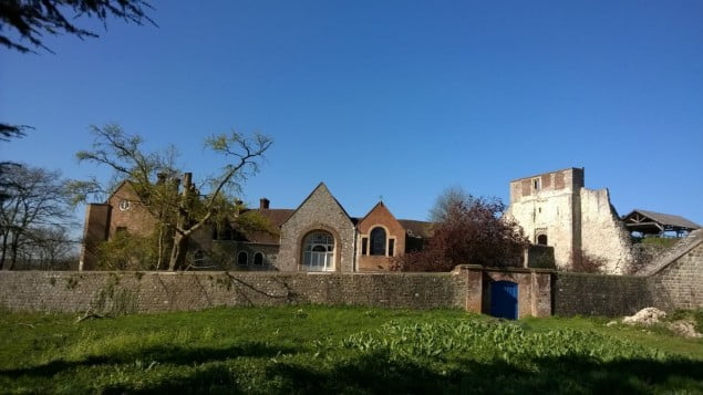 brick castle ruins to the right, newly built stone building on the left, stone wall in front, grass in foreground.