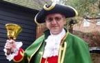 Town crier in green cloak, red waistcoat and tricorn hat, ringing bell.