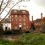 Large red brick building, river and grass surrounding.