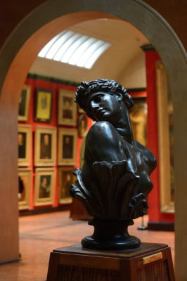 Bust of female head and shoulders on a plinth. Art gallery in the background