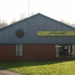Red brick building, with a blue roof, and green sign. Grass area in front and car park to the left.