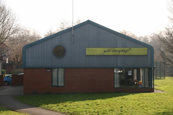 Red brick building, with a blue roof, and green sign. Grass area in front and car park to the left.