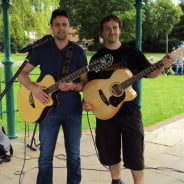 Two males with acoustic guitars standing under bandstand.