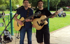 Two males with acoustic guitars standing under bandstand.