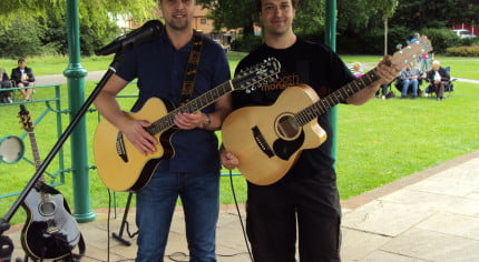 Two males with acoustic guitars standing under bandstand.