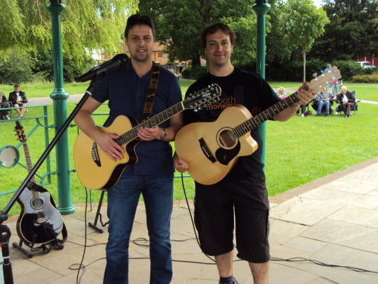 Two males with acoustic guitars standing under bandstand.