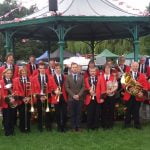 Group of male and female brass band members standing in front of band stand.
