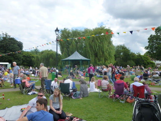 Groups of people, in the meadow, bandstand in background.