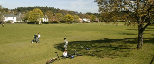 Three men playing football on village green. Large open green space with houses in foreground.