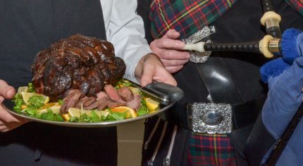 Chef holding venison joint on silver tray and piper holding bag pipes.