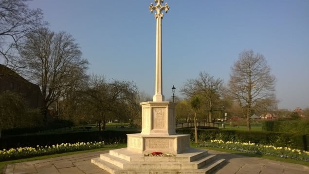 White stone war memorial. Paving at base. Yellow spring flowers in background. Blue sky. 