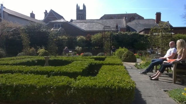 green hedges, footpaths, one male and one female sitting on a bench talking.