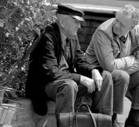 2 males sat on a wall, plants to the left. Black and white photo.