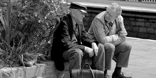 2 males sat on a wall, plants to the left. Black and white photo.