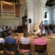 male playing guitar, audience sat on chairs in stone wall church. brass organ to the left.