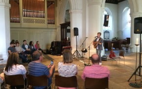 male playing guitar, audience sat on chairs in stone wall church. brass organ to the left.