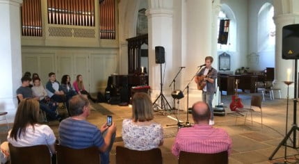 male playing guitar, audience sat on chairs in stone wall church. brass organ to the left.