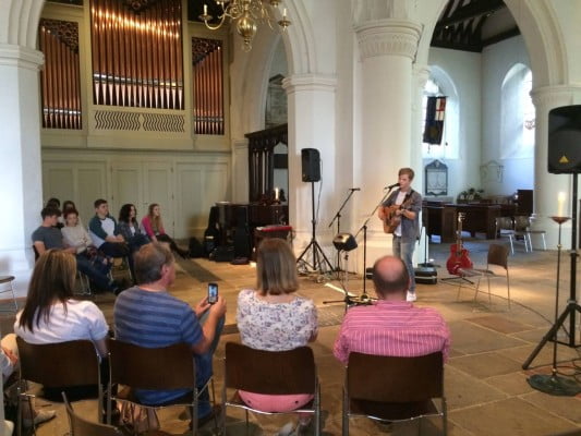 male playing guitar, audience sat on chairs in stone wall church. brass organ to the left.