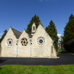 White stone chapel, tarmac driveway around, trees in the background.