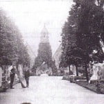 Old photo of cemetery, chapel in background, 2 males to the left.