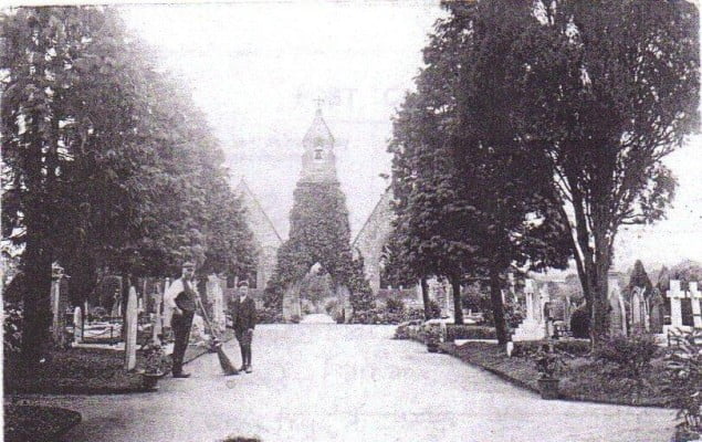 Old photo of cemetery, chapel in background, 2 males to the left.