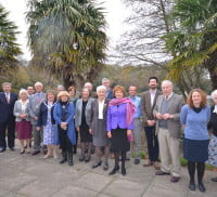 A group of adults standing in front of a flowerbed with trees behind.