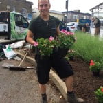 A male holding two plants in front of flower bed.