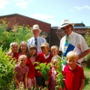 Two males with a group of school children in a garden.