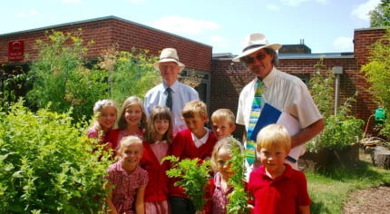 Two males with a group of school children in a garden.