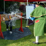 Male in stocks while male throws an bucket of water at him