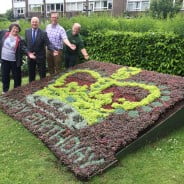 Three males and one female standing behind a flower bed in the design of a crown and with the words happy birthday.