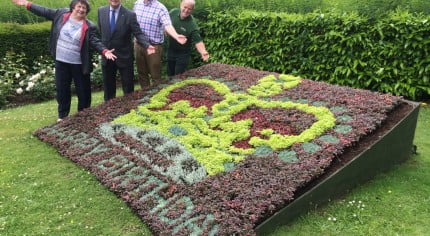 Three males and one female standing behind a flower bed in the design of a crown and with the words happy birthday.