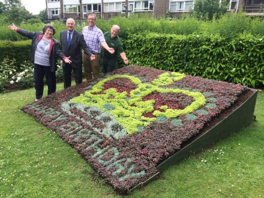 Three males and one female standing behind a flower bed in the design of a crown and with the words happy birthday.