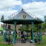 Three males playing electric guitar in a bandstand in a park.