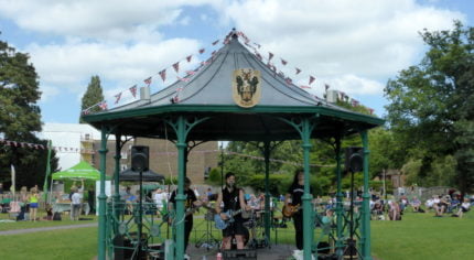 Three males playing electric guitar in a bandstand in a park.