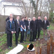 Four boys in school uniform with five adults. Standing in front of newly planted hedge.