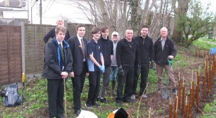 Four boys in school uniform with five adults. Standing in front of newly planted hedge.