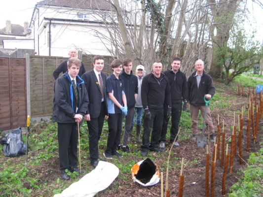 Four boys in school uniform with five adults. Standing in front of newly planted hedge.