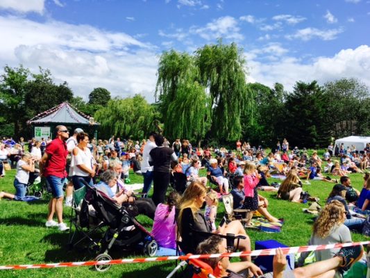 Large crowd of people sit and stand on grass to watch dance festival. Blue sky.