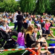 Large crowd of people sit and stand on grass to watch dance festival. Blue sky.