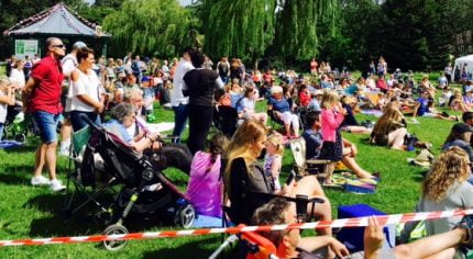 Large crowd of people sit and stand on grass to watch dance festival. Blue sky.