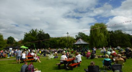 People sitting on grass and at picnic tables. Blue sky, bandstand in background.