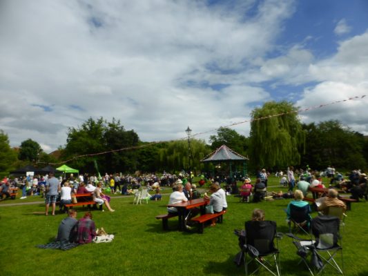 People sitting on grass and at picnic tables. Blue sky, bandstand in background.