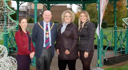 Mayor and three females in front of bandstand decorated with Christmas decorations.
