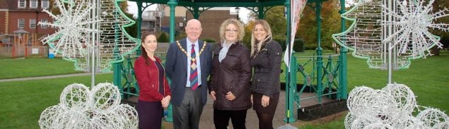 Mayor and three females in front of bandstand decorated with Christmas decorations.