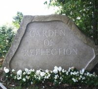 Large stone engraved with the words Garden of Reflection. White flowers at base of stone.