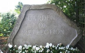 Large stone engraved with the words Garden of Reflection. White flowers at base of stone.