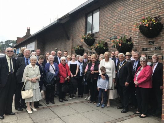 Large group of people standing in front of a wall displaying plaques and hanging baskets.