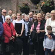 Large group of people standing in front of a wall displaying plaques and hanging baskets.