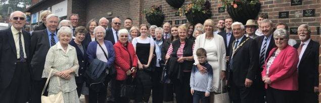 Large group of people standing in front of a wall displaying plaques and hanging baskets.